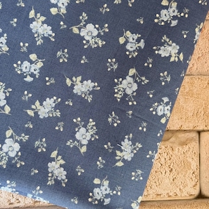 May include: A dark blue fabric featuring a repeating pattern of small, light blue and white floral clusters with delicate green leaves. The textile has a woven texture and is displayed in a folded orientation, showcasing the consistent floral print.