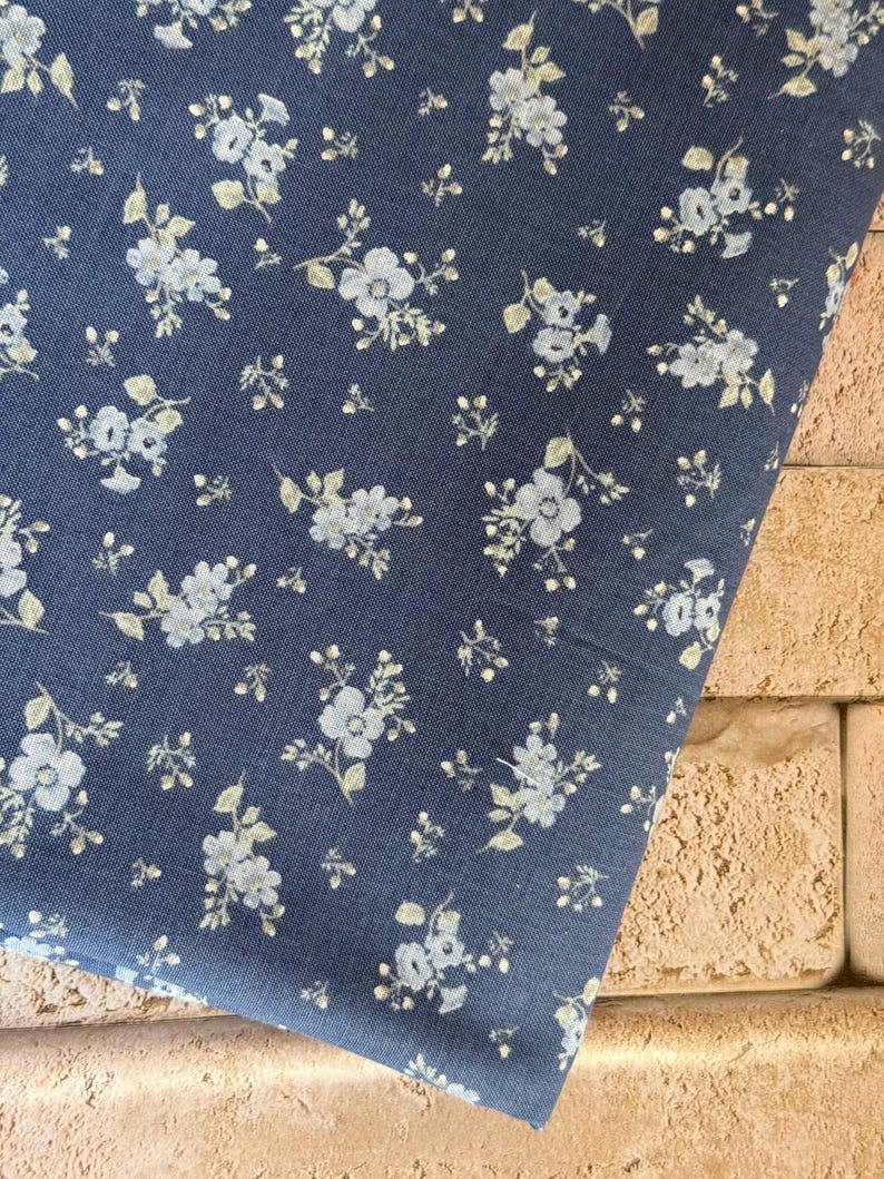 May include: A dark blue fabric featuring a repeating pattern of small, light blue and white floral clusters with delicate green leaves. The textile has a woven texture and is displayed in a folded orientation, showcasing the consistent floral print.