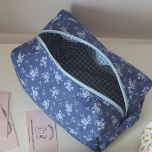 Könnte beinhalten: Ein gestepptes blaues Stoffetui mit weißem Blumenmuster und einem hellblauen Reißverschluss. Das Innere ist mit einem blau-weiß karierten Vichy-Stoff ausgekleidet. Eine kleine rechteckige Karte mit dem Text ARMESETSY ist zu sehen.