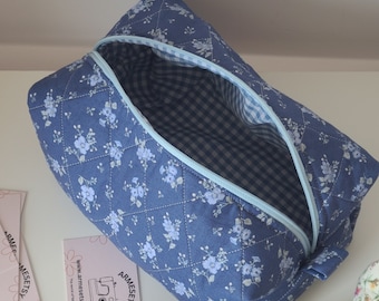 May include: A quilted blue fabric pouch featuring a white floral pattern and a light blue zipper. The interior is lined with a blue and white gingham check fabric. A small rectangular card with the text ARMESETSY and a sewing machine icon is visible.
