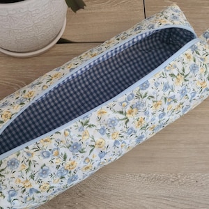 Peut inclure: Une pochette zippée en tissu blanc avec une doublure en vichy bleu et blanc. La pochette est décorée d'un motif floral dans les tons bleu, jaune et blanc.