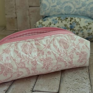 Puede incluir: Un estuche de tela acolchada con un estampado floral rosa sobre fondo blanco. El estuche tiene una cremallera rosa en el borde superior y un forro interior a cuadros rosas y blancos visible a trav&eacute;s de la abertura de la bolsa rectangular.