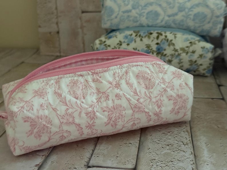Puede incluir: Un estuche de tela acolchada con un estampado floral rosa sobre fondo blanco. El estuche tiene una cremallera rosa en el borde superior y un forro interior a cuadros rosas y blancos visible a trav&eacute;s de la abertura de la bolsa rectangular.