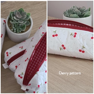 Puede incluir: Un estuche acolchado blanco con un patr&oacute;n de cerezas rojas y peque&ntilde;os puntos negros. El estuche tiene una cremallera roja y un forro interior de cuadros vichy rojos y blancos. El texto Cherry pattern aparece en blanco en la parte inferior.
