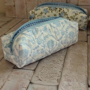 Puede incluir: Un estuche rectangular acolchado con un estampado floral en azul claro y blanco, con una cremallera azul a juego. La tela tiene una textura suave y pespunteada, y el dise&ntilde;o incluye un forro interior a cuadros en azul y blanco.