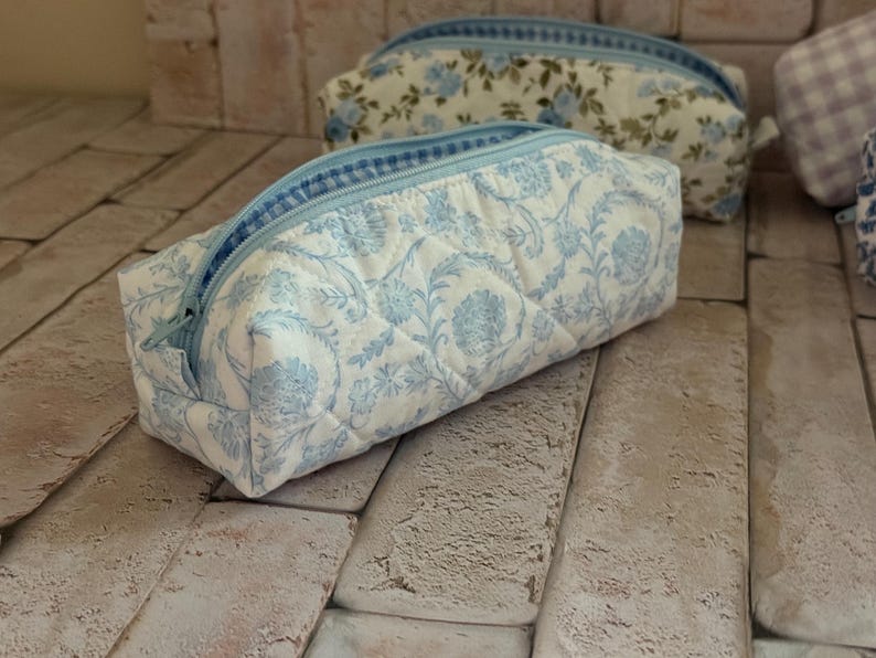 Puede incluir: Un estuche rectangular acolchado con un estampado floral en azul claro y blanco, con una cremallera azul a juego. La tela tiene una textura suave y pespunteada, y el dise&ntilde;o incluye un forro interior a cuadros en azul y blanco.