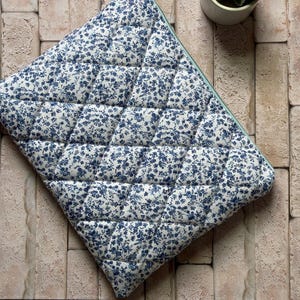 Könnte beinhalten: Eine rechteckige, gesteppte Laptoptasche mit weißem Stoffgrund und einem dichten blauen Blumenmuster. Die Tasche ist mit einer rautenförmigen Steppstruktur versehen und verfügt über einen Reißverschluss an der Oberkante für sicheren Verschluss.