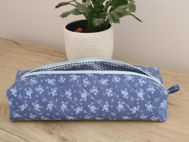 A blue quilted pencil case with a floral pattern sits on a wooden surface, with a potted plant in the background.