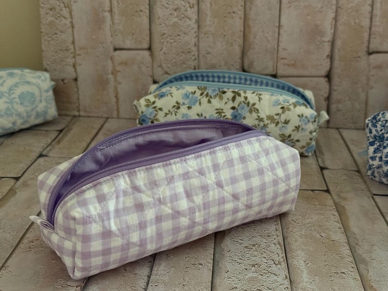Puede incluir: Un estuche acolchado con un patr&oacute;n de cuadros vichy en color violeta claro y blanco, con una cremallera violeta a juego y un forro interior liso del mismo tono. La bolsa rectangular tiene una textura suave y acolchada con cierre superior.