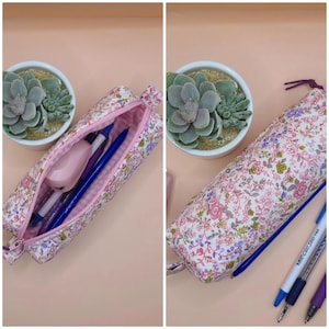 Puede incluir: Un estuche cil&iacute;ndrico de tela con un estampado floral en tonos rosa, verde y morado. El estuche se muestra abierto, revelando un interior rosa con bol&iacute;grafos y una grapadora, y cerrado, con tres bol&iacute;grafos colocados al lado.