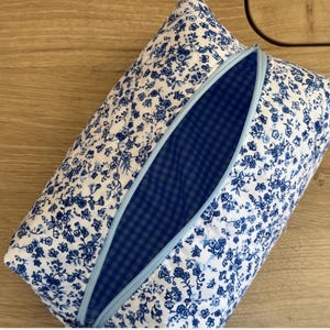 Peut inclure: Une trousse rectangulaire zippée présentant un extérieur en tissu blanc avec un motif floral bleu dense. La trousse possède une fermeture éclair bleu clair et une doublure intérieure en tissu à carreaux vichy bleu foncé et blanc.