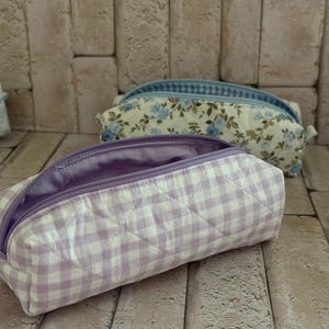 Puede incluir: Un estuche acolchado con un patr&oacute;n de cuadros vichy en color violeta claro y blanco, con una cremallera violeta a juego y un forro interior liso del mismo tono. La bolsa rectangular tiene una textura suave y acolchada con cierre superior.