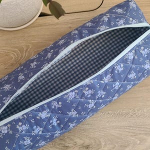 May include: A quilted fabric pouch in a dusty blue color featuring a white floral pattern. The exterior has a diamond-stitched texture, a light blue zipper closure, and a small fabric loop. The interior is lined with a black and white gingham fabric.