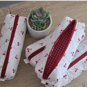 Puede incluir: Tres estuches acolchados blancos con un patr&oacute;n de cerezas rojas y cremalleras rojas. La tela tiene una textura de puntos sutil y un estuche est&aacute; abierto para revelar un forro interior de cuadros vichy rojos y blancos con una suculenta.