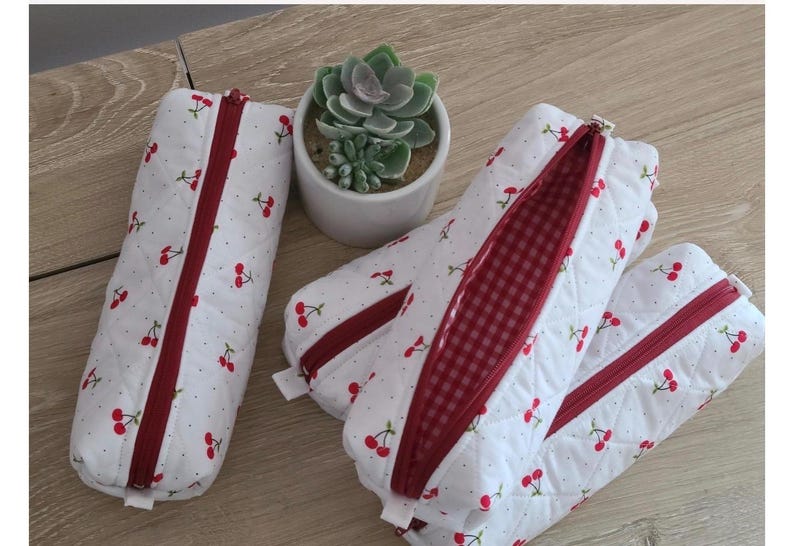 Puede incluir: Tres estuches acolchados blancos con un patr&oacute;n de cerezas rojas y cremalleras rojas. La tela tiene una textura de puntos sutil y un estuche est&aacute; abierto para revelar un forro interior de cuadros vichy rojos y blancos con una suculenta.