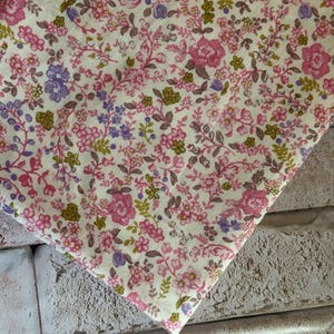 May include: A piece of fabric featuring a dense floral pattern on a cream background. The design consists of small pink roses, purple blossoms, and olive green leaves arranged in a repeating botanical print across the entire surface of the textile.