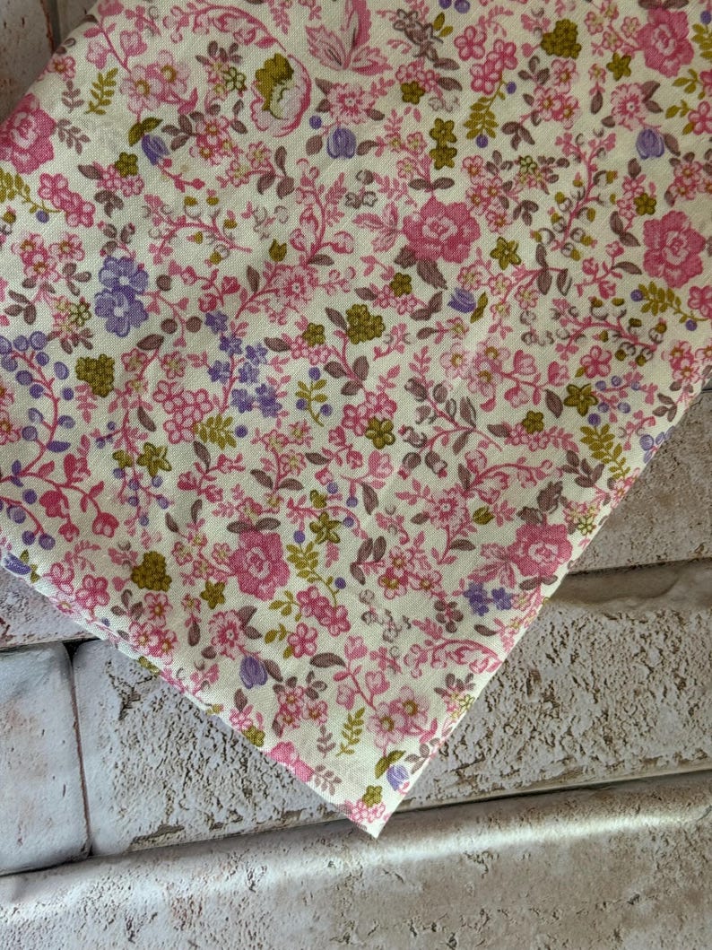 May include: A piece of fabric featuring a dense floral pattern on a cream background. The design consists of small pink roses, purple blossoms, and olive green leaves arranged in a repeating botanical print across the entire surface of the textile.