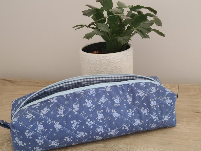 A blue quilted bag with a floral pattern sits on a wooden surface, with a potted plant in the background.
