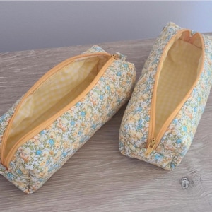 Puede incluir: Dos estuches rectangulares con un estampado floral de peque&ntilde;as flores azules, amarillas y verdes sobre un fondo crema. Cada estuche tiene una cremallera amarilla y un forro interior de tela de cuadros vichy en color amarillo y blanco.