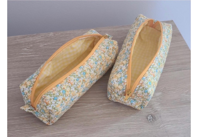 Puede incluir: Dos estuches rectangulares con un estampado floral de peque&ntilde;as flores azules, amarillas y verdes sobre un fondo crema. Cada estuche tiene una cremallera amarilla y un forro interior de tela de cuadros vichy en color amarillo y blanco.