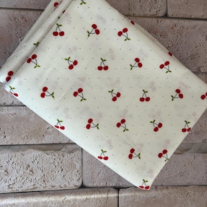 May include: A folded piece of off-white fabric featuring a repeating pattern of small red cherries with green stems and leaves, interspersed with tiny black dots across the entire surface of the textile material.