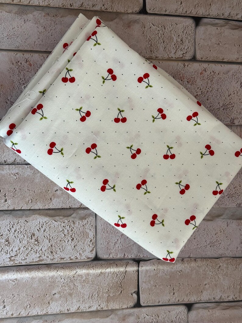 May include: A folded piece of off-white fabric featuring a repeating pattern of small red cherries with green stems and leaves, interspersed with tiny black dots across the entire surface of the textile material.