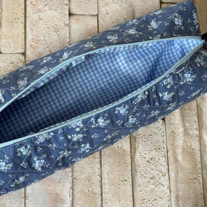 May include: A quilted fabric pencil case featuring a blue floral pattern on the exterior with a diamond-stitched texture. The interior is lined with a light blue and white gingham check fabric, and the case includes a light blue zipper closure.