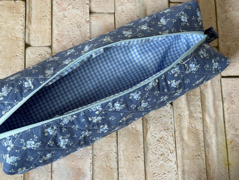 May include: A quilted fabric pencil case featuring a blue floral pattern on the exterior with a diamond-stitched texture. The interior is lined with a light blue and white gingham check fabric, and the case includes a light blue zipper closure.