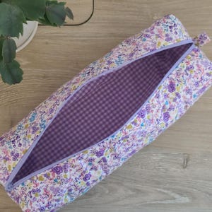 A floral patterned pencil case with a purple lining is placed on a wooden surface next to a potted plant.