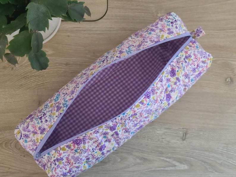A floral patterned pencil case with a purple lining is placed on a wooden surface next to a potted plant.