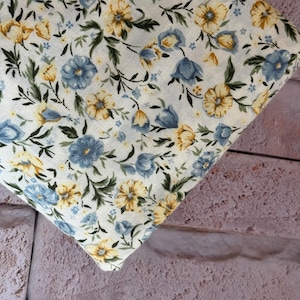 May include: A piece of off-white fabric featuring a repeating floral pattern. The design consists of small blue and yellow flowers with green leaves and stems scattered across the surface, creating a dense botanical print on the textile.