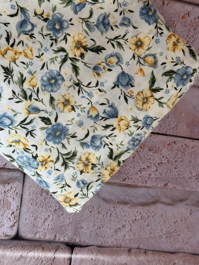May include: A piece of off-white fabric featuring a repeating floral pattern. The design consists of small blue and yellow flowers with green leaves and stems scattered across the surface, creating a dense botanical print on the textile.