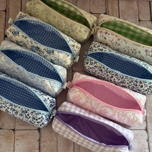 Puede incluir: Una colecci&oacute;n de siete estuches rectangulares de tela con varios estampados florales y de cuadros vichy. Cada estuche tiene una abertura con cremallera que revela forros interiores en contraste de telas a cuadros o lisas en verde, azul, rosa y morado.