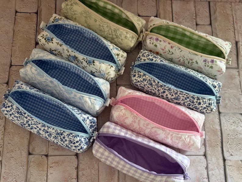 Puede incluir: Una colecci&oacute;n de siete estuches rectangulares de tela con varios estampados florales y de cuadros vichy. Cada estuche tiene una abertura con cremallera que revela forros interiores en contraste de telas a cuadros o lisas en verde, azul, rosa y morado.