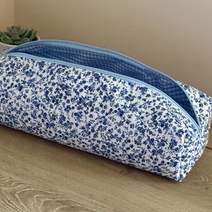 May include: A rectangular zipped pouch featuring a white fabric base with a dense blue floral pattern. The interior is lined with a blue and white gingham check fabric. The pouch has a light blue zip closure along the top edge.