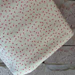 May include: A piece of light cream-colored fabric featuring a repeating pattern of small, dark pink tulip-like floral motifs. The textile has a visible woven texture and is folded, displaying the consistent print across the entire visible surface area.