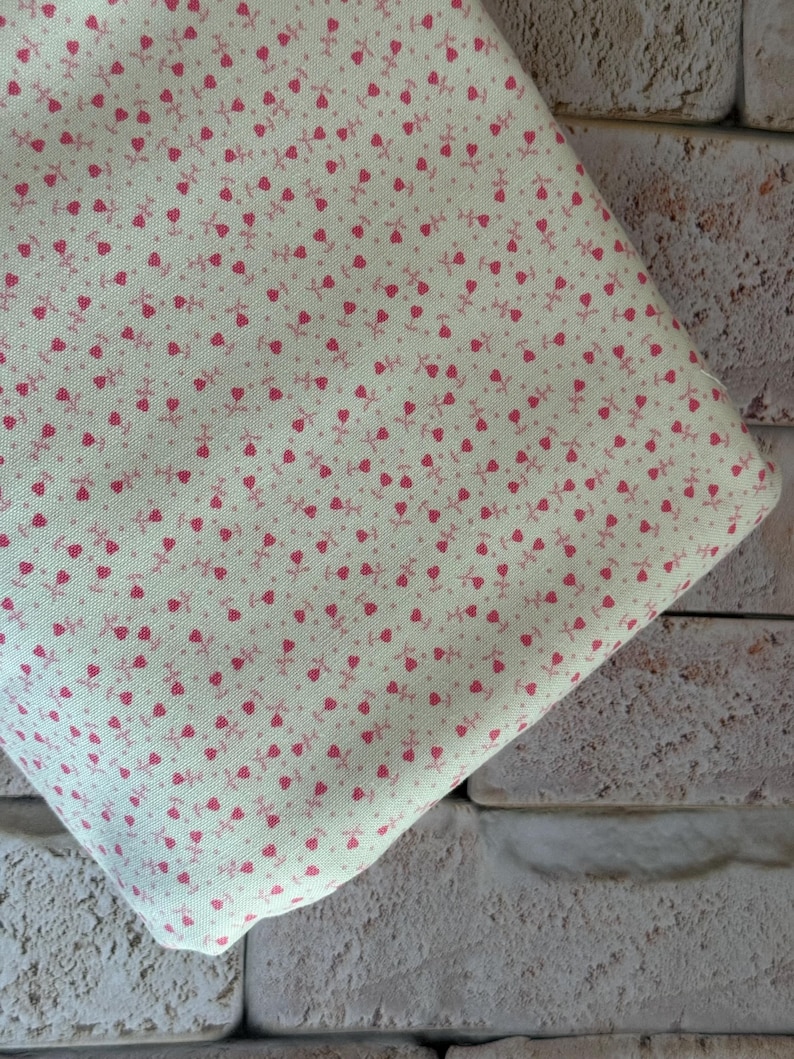 May include: A piece of light cream-colored fabric featuring a repeating pattern of small, dark pink tulip-like floral motifs. The textile has a visible woven texture and is folded, displaying the consistent print across the entire visible surface area.