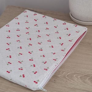 A white laptop bag with a red and white cherry pattern is placed on a wooden surface next to a white ceramic planter.