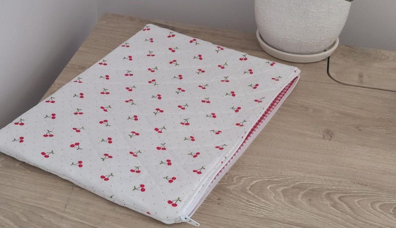 A white laptop bag with a red and white cherry pattern is placed on a wooden surface next to a white ceramic planter.