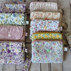 May include: A collection of small rectangular fabric pouches featuring various floral and dotted patterns in pastel and vibrant colors. Each pouch includes a front flap with a snap button closure, a metal ring attachment, and a matching fabric wrist strap.