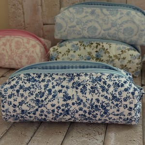 Puede incluir: Una colecci&oacute;n de estuches rectangulares de tela con varios estampados florales en tonos azul, rosa y blanco. Cada estuche tiene un cierre de cremallera y una forma estructurada, destacando uno en primer plano con un denso estampado floral azul.