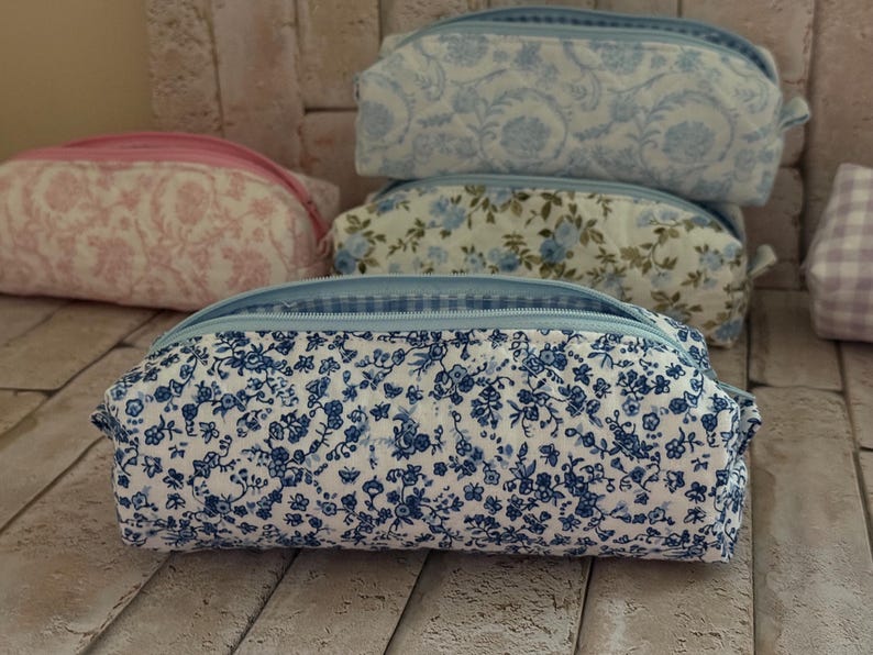 Puede incluir: Una colecci&oacute;n de estuches rectangulares de tela con varios estampados florales en tonos azul, rosa y blanco. Cada estuche tiene un cierre de cremallera y una forma estructurada, destacando uno en primer plano con un denso estampado floral azul.