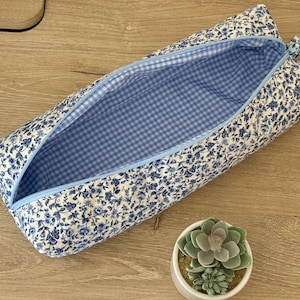 May include: A rectangular fabric pencil case featuring a blue and white floral pattern on the exterior and a light blue and white gingham check lining. The case has a light blue zipper closure along the top edge and a soft, padded structure.