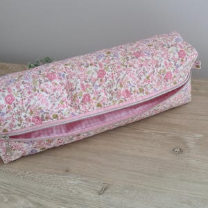 May include: A cylindrical fabric case featuring a floral pattern with pink, white, and green tones. The exterior is quilted, and the case includes a silver-toned metal zipper closure that reveals a light pink interior lining with a subtle checkered pattern.