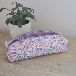 May include: A floral print pencil case with a lavender zipper and gingham lining. The case is open, revealing the interior. A potted plant sits in the background on a wooden surface.