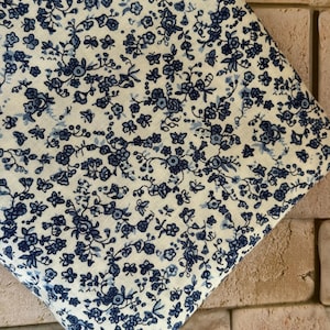 May include: A piece of fabric featuring a dense, repeating pattern of small dark blue flowers and delicate leaves set against a light cream-colored background. The textile displays a consistent floral motif across its entire visible surface area.