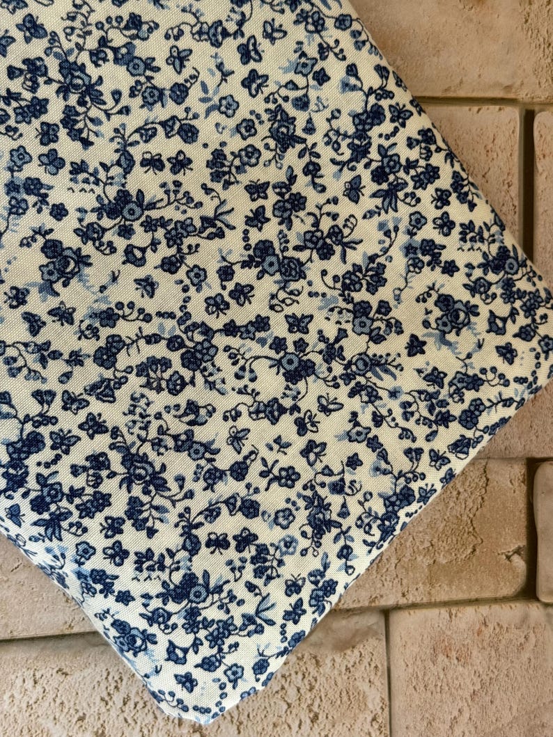 May include: A piece of fabric featuring a dense, repeating pattern of small dark blue flowers and delicate leaves set against a light cream-colored background. The textile displays a consistent floral motif across its entire visible surface area.