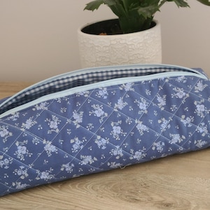A blue quilted pencil case with a floral pattern sits on a wooden surface, with a potted plant in the background.