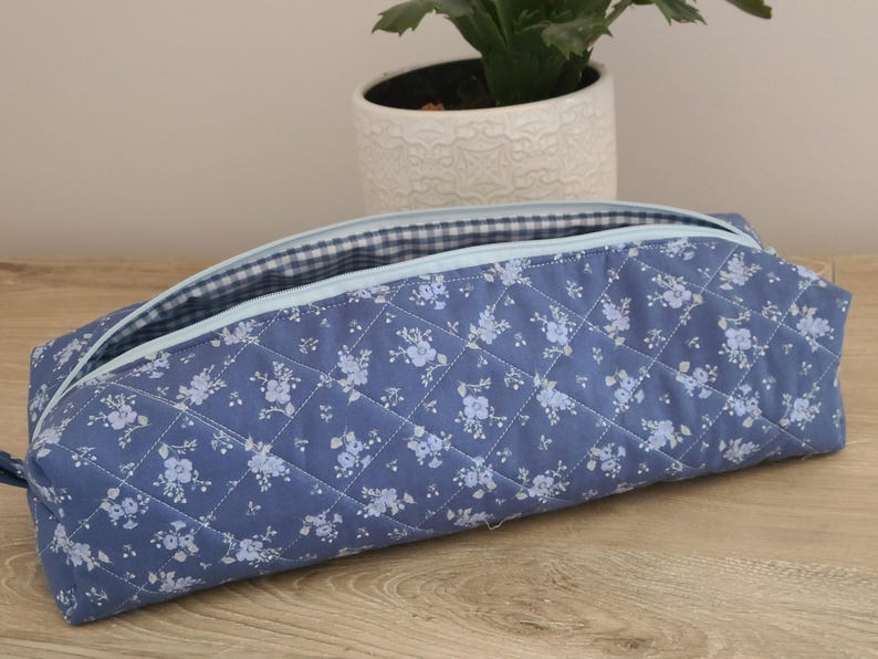 A blue quilted pencil case with a floral pattern sits on a wooden surface, with a potted plant in the background.