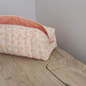 May include: A rectangular quilted zippered pouch featuring a light cream fabric with a repeating pattern of small peach-colored flowers. The interior is lined with a bright orange and white gingham check fabric, and the pouch is partially open.